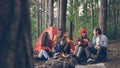 Joyful tourists men and women are sharing stories sitting around campfire and laughing getting warm near burning flame