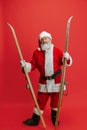 Full-length portrait of senior man wearing Santa Claus costume, holding skis isolated over red background. Winter Royalty Free Stock Photo