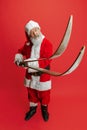 Full-length portrait of senior man wearing Santa Claus costume, holding skis  over red background. Winter Royalty Free Stock Photo