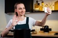 Joyful woman in an apron cooks at home and tastes a dish, shooting a video tutorial on the phone in the kitchen interior Royalty Free Stock Photo