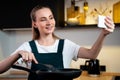 Joyful woman in an apron cooks at home and tastes a dish, shooting a video tutorial on the phone in the kitchen interior Royalty Free Stock Photo