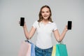 joyful woman holding smartphones with blank screen and shopping bags on grey, black friday concept.