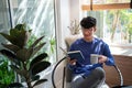 Joyful young Asian man in casual clothes and eyeglasses reading book while relaxing at living room of his home Royalty Free Stock Photo