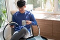 Joyful young Asian man in casual clothes and eyeglasses reading book while relaxing at living room of his home Royalty Free Stock Photo