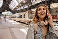 Joyful young caucasian woman talking on phone, waiting for train standing at station. Royalty Free Stock Photo