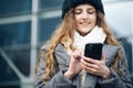 Joyful young female in good mood typing and scrolling on smartphone outdoors. Winter concept. Female using smartphone