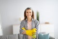 Joyful young housemaid in rubber gloves holding detergent and smiling at camera at home. Sanitary service concept