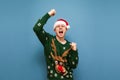 Joyful young man in santa hat and warm christmas sweater stands against a blue background and rejoices in victory. Happy man won,
