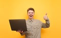 Joyful young man in shirt and glasses stands with laptop in hands on yellow background, looks into camera and shows thumb up. Royalty Free Stock Photo