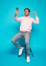 joyful young man sitting on chair and holding mobile phone isolated over light blue, celebrating financial success Royalty Free Stock Photo