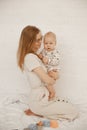Joyful young mother sit on lap on bed and hold little baby on white background. Home family photo of woman and cute Royalty Free Stock Photo