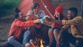 Joyful young people friends are clinking glasses with drinks sitting around fire in forest with warm marshmallow on Royalty Free Stock Photo