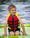 joyful young smiling boy kneels on floating mat, wearing lifejacket as he plays in lake water Royalty Free Stock Photo