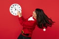 Joyful young woman dressed in Christmas santa sweater pointing finger on white big clock over red background. Royalty Free Stock Photo