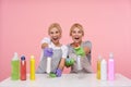 Joyous young lovely blonde cleaning ladies looking happily at camera with wide smiles while posing over pink background with spray