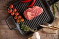 Juicy, aged raw ribeye steak in a grill pan with red chili peppers, rosemary, red tomatoes on a branch, a head of garlic, burlap, Royalty Free Stock Photo