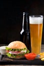 Juicy burger, french fries, sauce, beer in a bottle and glass on a dark board and wooden background.Fast food Royalty Free Stock Photo