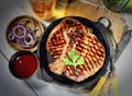 Juicy steak with parsley in a frying pan, onions, beer, vintage knife and fork, a napkin on a gray background Royalty Free Stock Photo