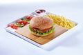 Juicy tasty hamburger on a wooden cutting board with french fried fries, vegetables and ketchup. Isolated composition on Royalty Free Stock Photo