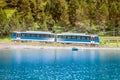 Train passing through Nuria valley against foreground of blue mountain lake, Spain Pyrenees Royalty Free Stock Photo