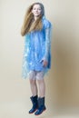 Jumping Caucasian Teenager Girl In Blue Dress With Transparent Raincoat and Rubber Boots Against Beige Background