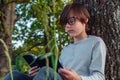 Attentive boy with square glasses holding student book in hands and reading while sitting leaning on tree in park.