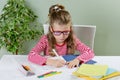 A junior schoolgirl with glasses writes something with her left hand in the notebook and sits at the table. The child is left-hand Royalty Free Stock Photo