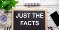 JUST THE FACTS written on a black background near pencils, a smartphone, a white notepad and a green plant in a pot Royalty Free Stock Photo