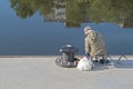 Old fisherman on city quay with three fishing rods on background of a river with houses and trees reflected in it.