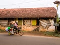 An Indian man rides a bicycle past a rustic building with slanted roofs in a