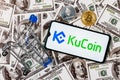 A smartphone with the KuCoin logo, a shopping cart and a cryptocoins on the dollar bills