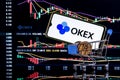 A smartphone with the OKEx logo in shopping cart on the background of the live trading webpage