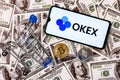 A smartphone with the OKEx logo, a shopping cart and a cryptocoins on the dollar bills