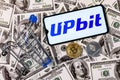 A smartphone with the Upbit logo, a shopping cart and a cryptocoins on the dollar bills