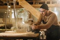 Keen carpenter in watch cap shaving wood with electric plane in a big workshop. Royalty Free Stock Photo