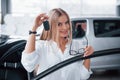 With keys in hand. Cute girl in eyewear stands near the car in auto saloon. Probably her next purchase Royalty Free Stock Photo
