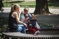 A young mother with a baby in a backpack and a friend are relaxing on a bench in a city park