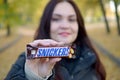 KHARKOV, UKRAINE - OCTOBER 8, 2019: A young caucasian brunette girl shows Snickers chocolate bar in autumn park. Snickers