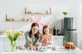 Kid and attractive mother with bunny ears near chicken eggs, decorative rabbits, easter bread and tulips Royalty Free Stock Photo