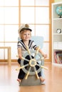 Kid boy dressed as a captain or sailor plays on chair like ship in his room. Child rotates the wooden steering wheel. Royalty Free Stock Photo