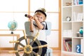 Kid boy dressed captain or sailor plays on chair as ship in his room. Child looks through telescope.