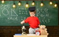 Kid boy in graduate cap near microscope in classroom, chalkboard on background. First former interested in studying