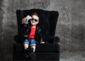 Kid boy millionaire in leather jacket and red t-shirt is sitting in big luxury armchair handing us a bundle of dollars cash