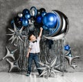 Kid boy stands on background of silver and blue metallic balloons for birthday party holding hand up and smiles Royalty Free Stock Photo