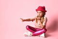 Kid in casual outfit is looking surprised, pointing by forefinger at something, sitting on floor with crossed legs against pink Royalty Free Stock Photo