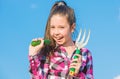 Kid cheerful gardener holds rake and cucumber blue sky background. Girl gardener with hand rake. Gardening and