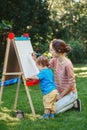 Kid girl with older sister, teacher standing outside in summer autumn park drawing on easel with markers, playing studying Royalty Free Stock Photo