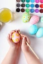 Kid hand painted Easter eggs, paints and brushes on a white table. Preparation for the holiday. Girls hands draw a pattern