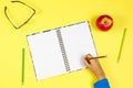 Kid hand writing on notebook, colorful pencils, glasses and fresh apple on yellow background