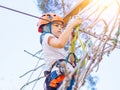 Kid in orange helmet climbing in trees on forest adventure park. Girl walk on rope cables and high suspension bridge in